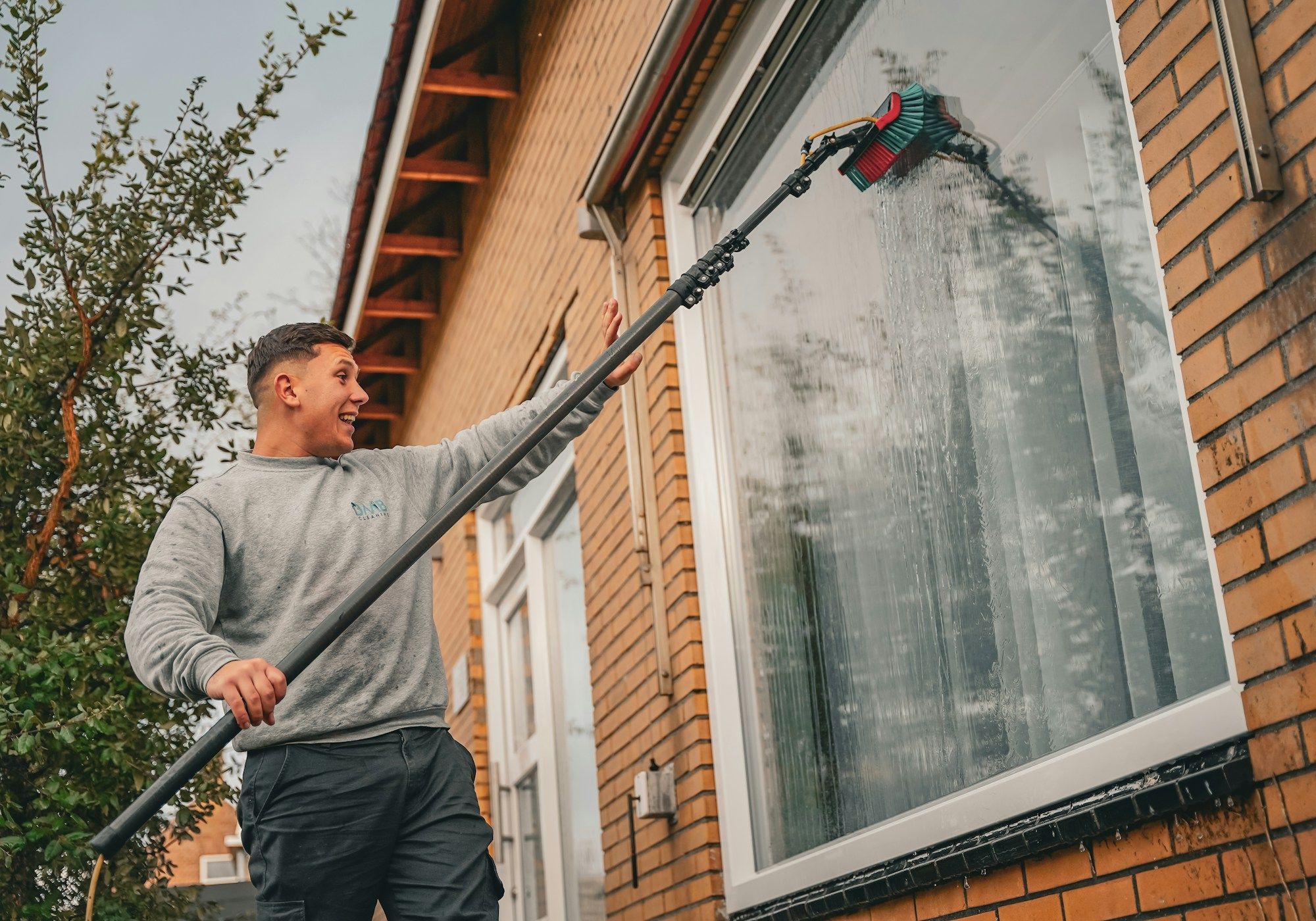 A window cleaner rinsing the front window of a brick house with a water-fed pole from the ground.
